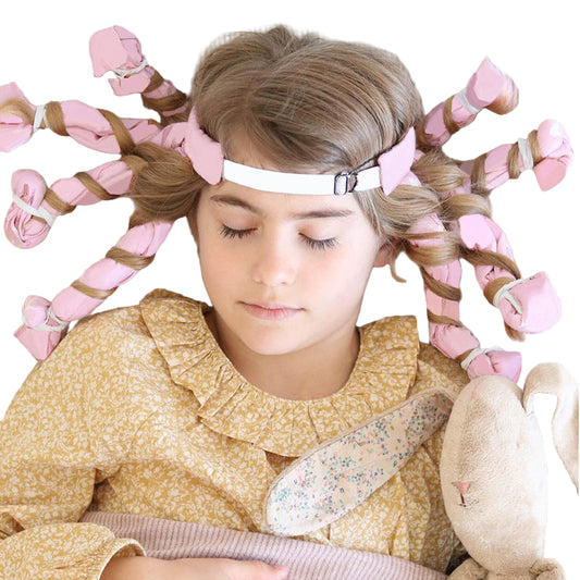 Young girl with braided hair and pink ribbons, wearing a yellow patterned dress, lying on a bed with a plush toy.